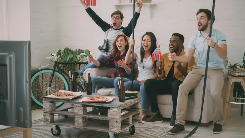 How to Host a Cricket Match - Excited friends cheering while watching a sports game on TV, holding flags and snacks.